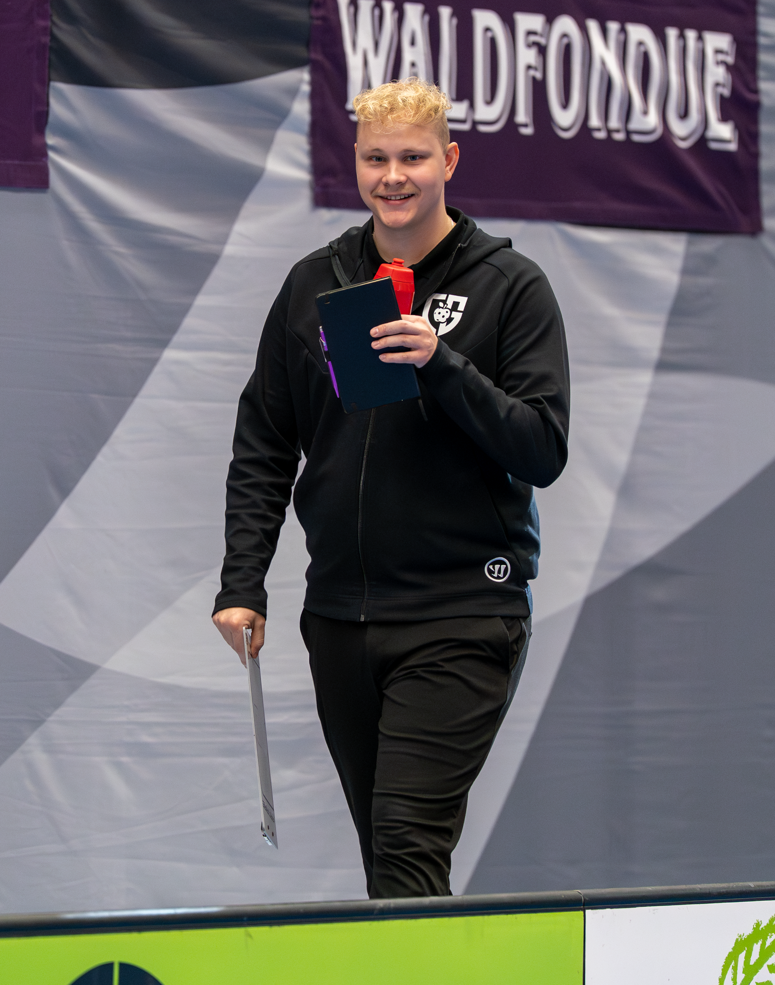Portrait of the personal football coach standing indoors in front of a banner, smiling at the camera and holding a clipboard and water bottle, wearing black sportswear.